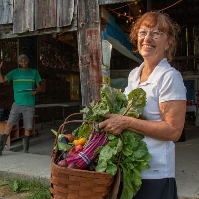 Woman leaving farm store with a basket full of fresh vegetables such as bright purple beets with green tops, bright red and orange peppers, red big tomatoes and curly green kale.