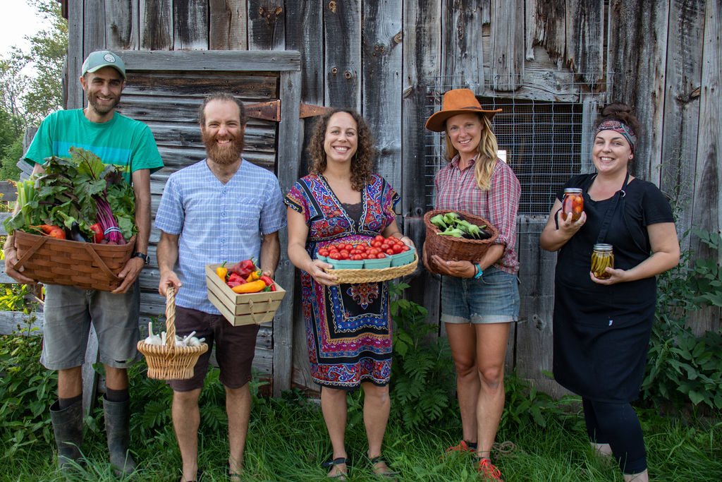 Farmers in front of barn holding many sized baskets full of fresh veggies, garlic and preserves